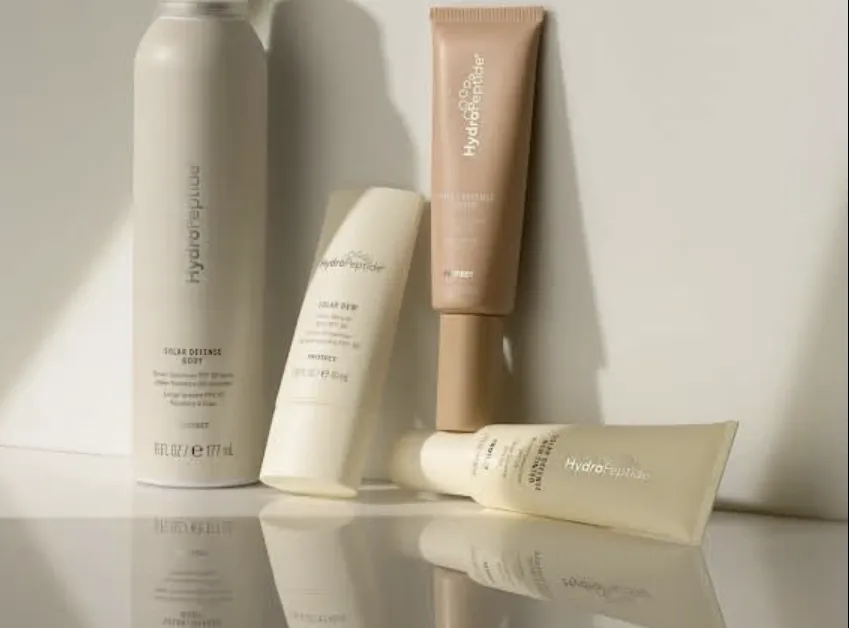 Cream-coloured HydroPeptide sun care bottles and tubes against a wall